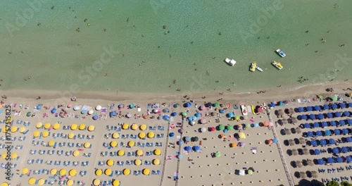 Aerial perpendicular view of a beach resort on a sandy beach. The sea is crystal clear. The beach club has yellow umbrellas, blue loungers, and vacationers sunbathing.