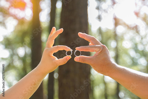 A couple holding up their two wedding rings against a sunlit, blurred green forest background.