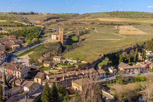 A picturesque view overlooks the Segovia Templar Church of Vera Cruz in a hilly valley, surrounded by a small village of brown-roofed houses below