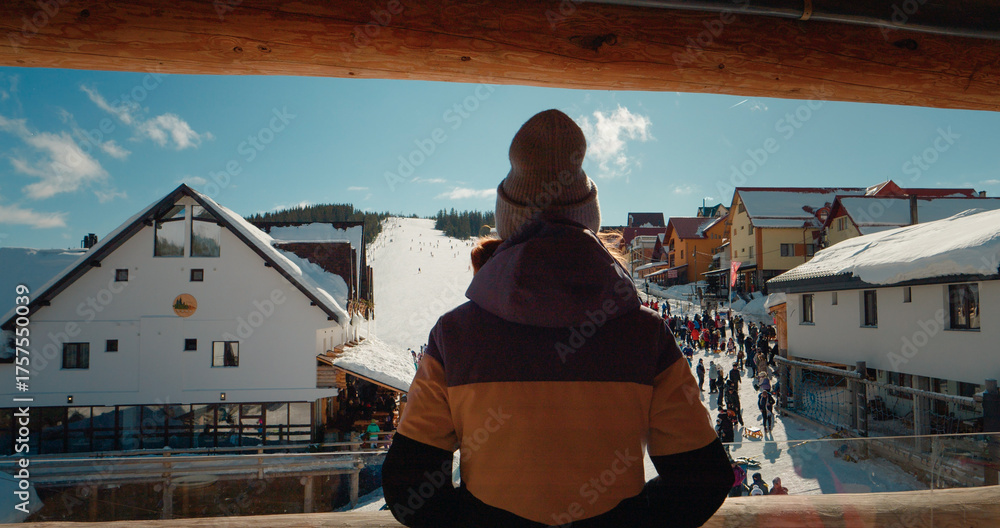 Fototapeta premium View from the hotel balcony to the snowing ski path resort. Back view of woman dancing while standing on winter resort balcony looking the tourists.