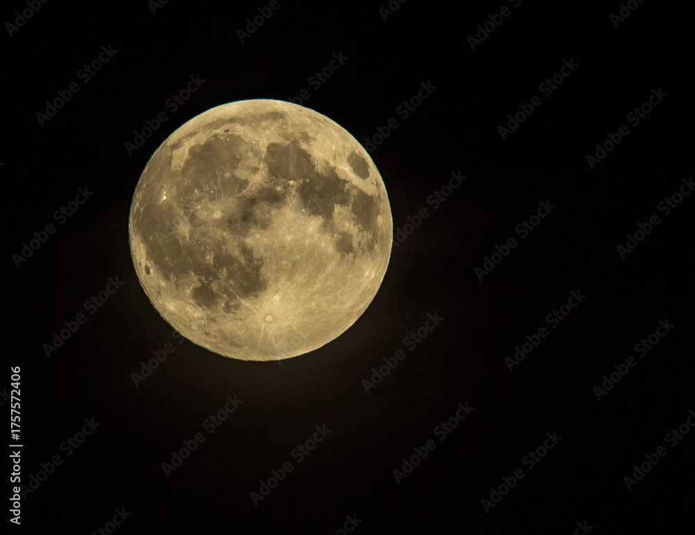 Obraz premium A detailed image of a full or nearly full Moon, illuminated by a yellowish glow, against a pitch-black night sky. Craters and lunar maria are clearly visible. 