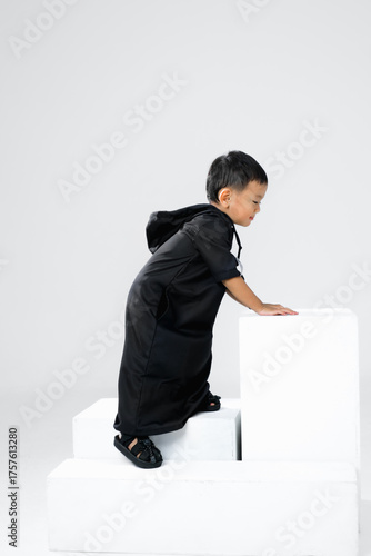 Little boy climbing white blocks in studio while wearing black thobe. Concept of playful Islamic kid, modern fashion, and active childhood lifestyle.