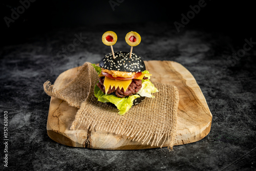 Close-up of monster-style hamburger with black bun, cheese teeth, bacon, and edible eyes on burlap over wooden board, top view food photography for Halloween