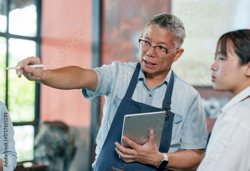 Photography Elderly asian male cafe owner in apron holding tablet while instructing employee