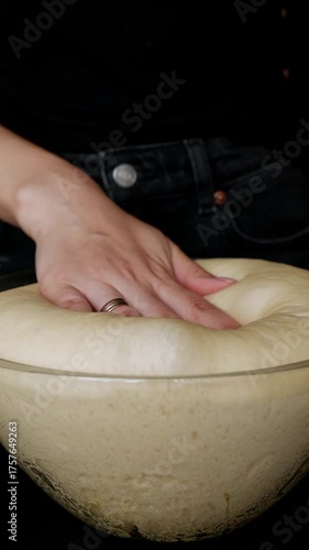 A woman’s hand presses down on soft, airy yeast dough in a glass bowl. Preparing homemade baked goods, baking bread, and letting dough rise. Concept of home cooking, fermentation, and artisan baking.