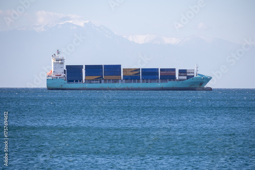 turquoise container ship sailing on a vast blue sea, carrying freight under a clear sky with a dramatic background of snow-capped, hazy mountains.