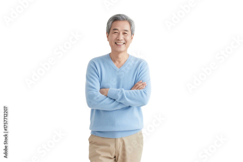 An elderly man in a light blue sweater stands with arms crossed, smiling in a simple background.