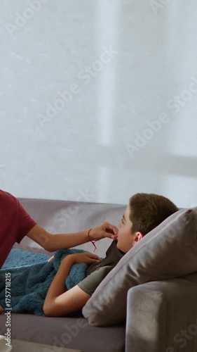 Caring sister checks thermometer used by sick brother resting under blue blanket on couch during quiet indoor moment filled with warmth empathy love and family support in natural daylight environment