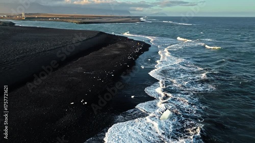 Aerial of coastline Diamond Beach Iceland. The drone moves above ocean waves meeting black volcanic sand and floating ice.