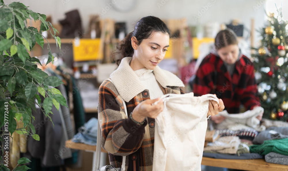 Obraz premium Armenian woman stands in a store against the background of a Christmas tree and chooses warm pants for the winter holidays. Buyer on sale before Christmas and New Year, holiday atmosphere around