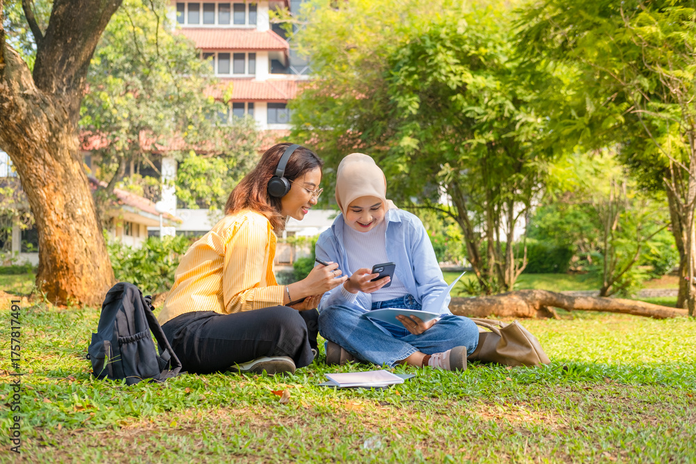 Fototapeta premium Two young woman while listening to audio lessons or podcasts on her laptop. Captured in a bright outdoor park near a university building.