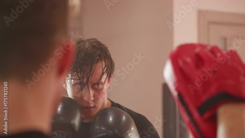 Focused boxer wearing black gloves trains with coach using red pads during intense sparring session, sweat dripping from face, eyes locked on target, demonstrating endurance, discipline in boxing gym
