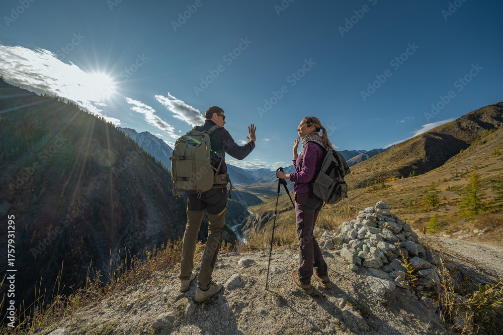 Fototapeta premium Travelers with backpacks stand on a mountain slope and clap their hands, with an autumn forest and a blue sky in the background. The admiration of victorious people. Achieving a goal in the mountains.