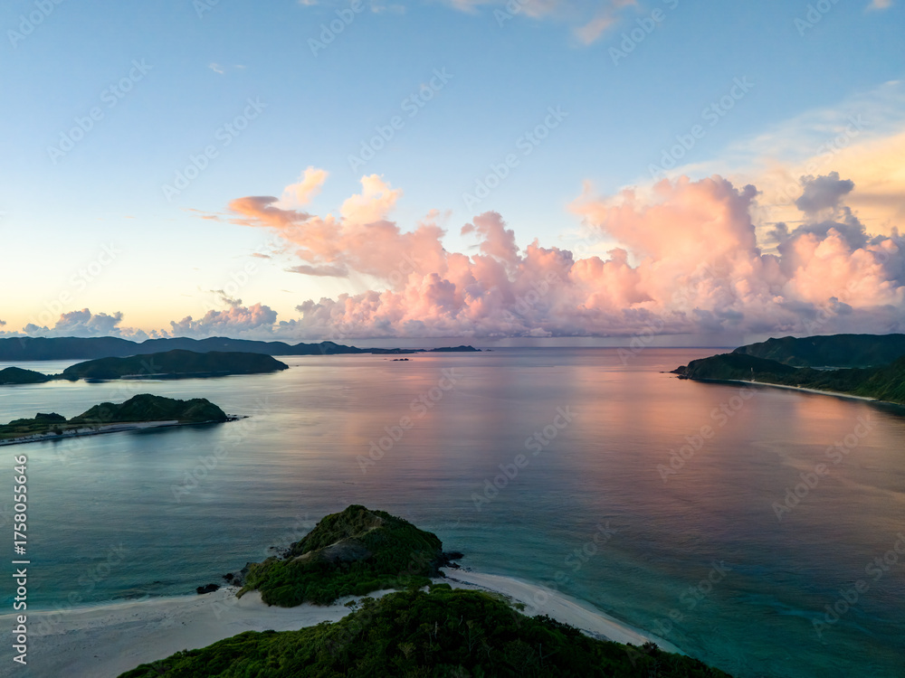 Fototapeta premium 座間味港で慶良間諸島の日の出をドローンで空中撮影している。 座間味島座間味港 沖縄県島尻郡慶良間諸島-2025 沖縄本島から40km西方にある諸島。 大小30以上の島々からなる島嶼群は全体が国立公園に指定されている。 珊瑚礁の海、白い砂浜、緑の山地は世界屈指の美しさ。 多くの貴重な動植物が生息している。 