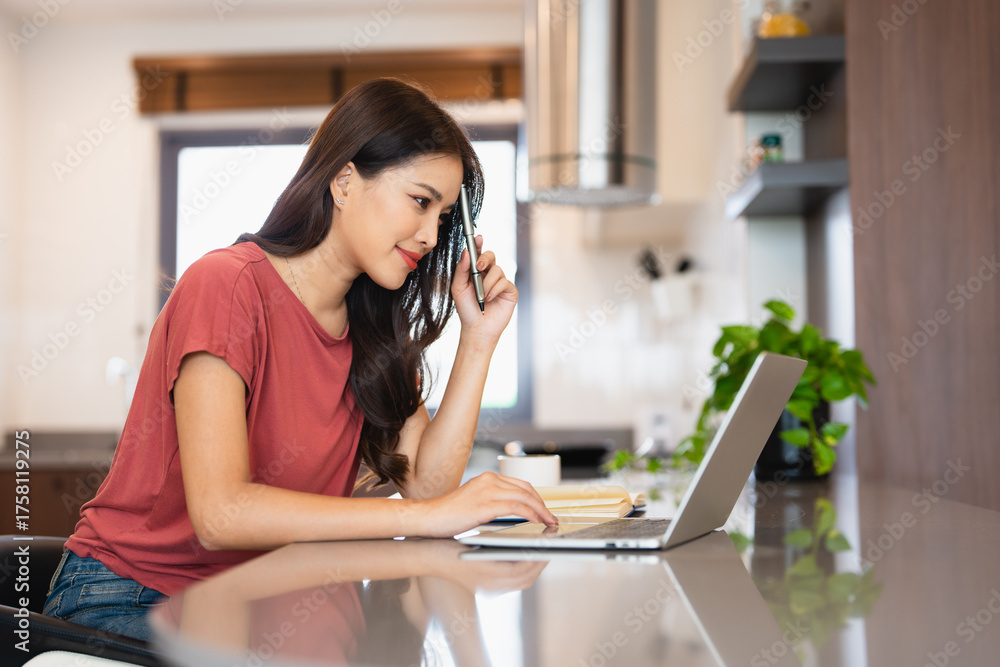 Obraz premium Young woman sitting in kitchen and working on laptop in weekend morning. Female using laptop on dining table .