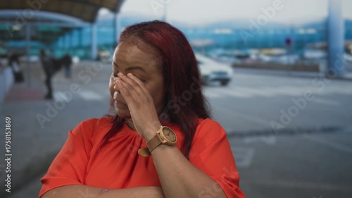Woman in red shirt pinching nose by airport terminal entrance while wearing wooden watch on wrist; disgust.