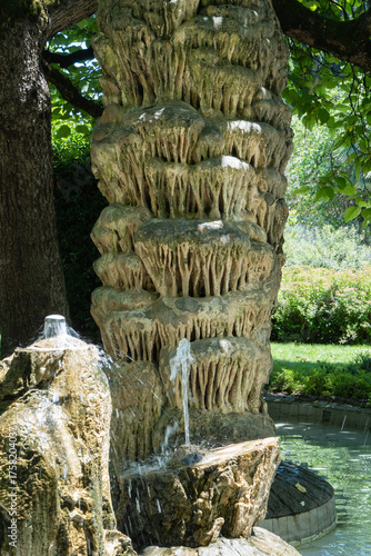 Unique stone fountain made of natural stalagmites, surrounded by lush greenery and tall trees, creating calm, natural atmosphere. Sochi City Park.