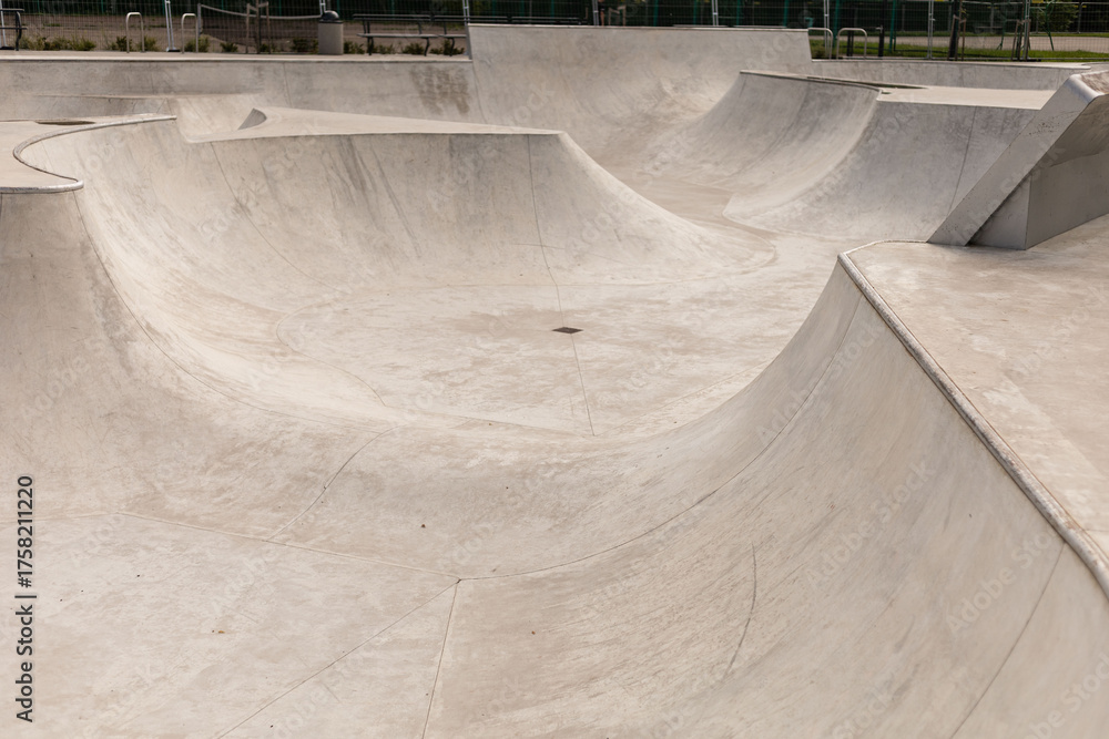 Fototapeta premium Multiple smooth concrete ramps and bowls are designed for skateboarding and BMX riding at a community skatepark in bright daylight. The empty park invites skaters to enjoy the space