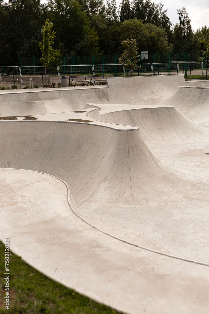 Fototapeta premium A modern skatepark features smooth, flowing concrete structures with various ramps and bowls, ideal for skateboarding and BMX activities. The park is surrounded by green trees and is well-maintained