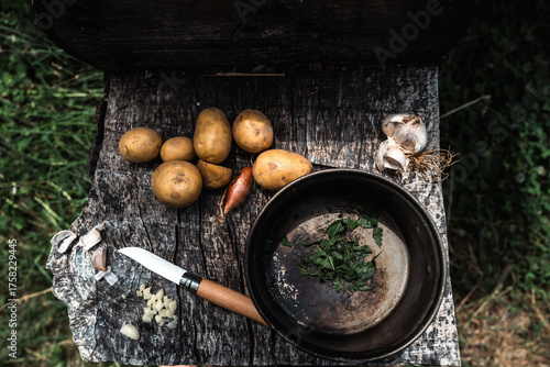Pommes de terre, ail, échalote et herbes fraîches disposés sur une planche en bois avec un couteau et une casserole. Scène rustique et naturelle de cuisine en plein air, évoquant la simplicité 
