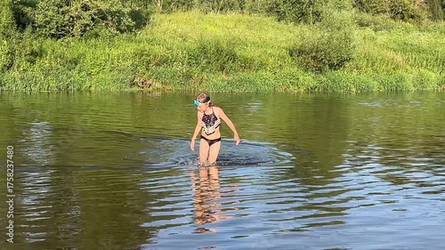 A girl in a black two-piece swimsuit goes in the the river.