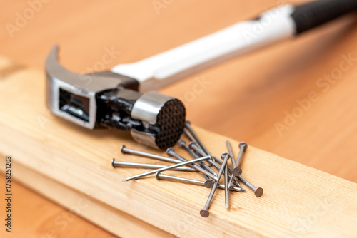 New modern hammer with a white handle lying on a wooden board next to a pile of steel nails for carpentry or repair work, close-up