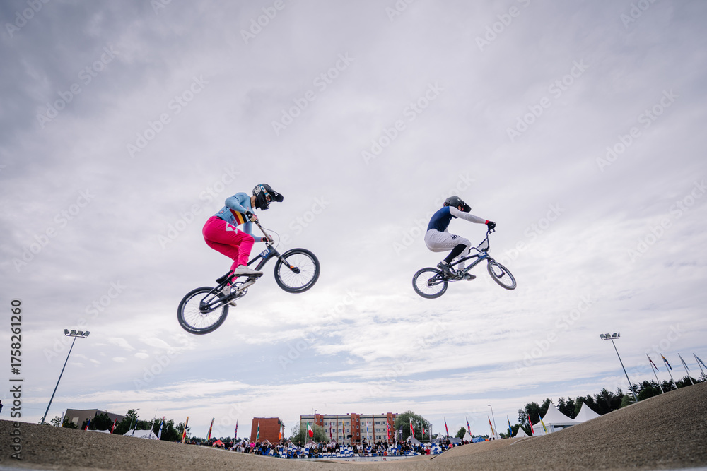 Fototapeta premium Valmiera, Latvia - July 12, 2025: BMX riders performing tricks in mid-air during competition with cloudy sky backdrop. BMX European championships
