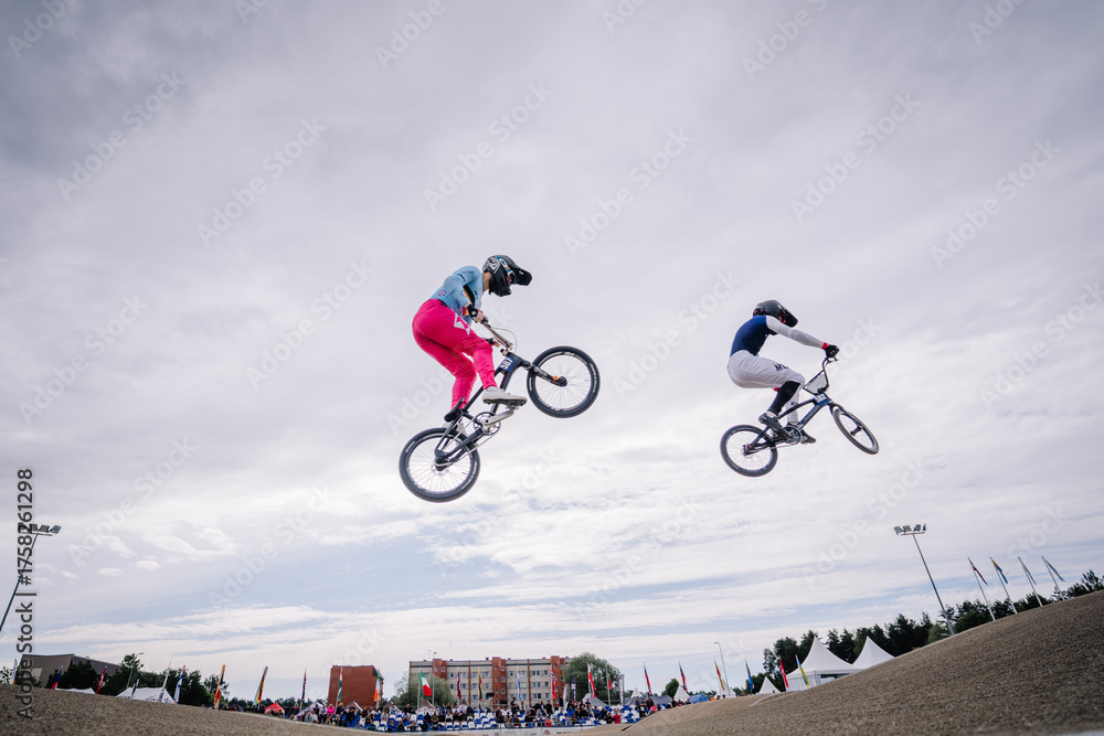 Fototapeta premium Valmiera, Latvia - July 12, 2025: BMX riders performing tricks in mid-air during competition with cloudy sky backdrop. BMX European championships
