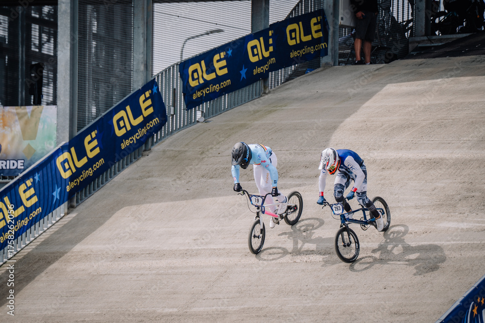 Fototapeta premium Valmiera, Latvia - July 12, 2025: Two male BMX riders racing on dirt track with ALE branding in background. BMX European championships