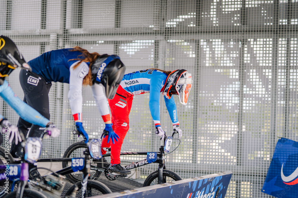 Fototapeta premium Valmiera, Latvia - July 12, 2025: BMX racers poised at starting gate ready for competition in vibrant athletic gear. BMX European championships