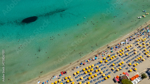 Aerial view of a beach resort on a sandy beach. The sea is crystal clear and emerald green. The beach club has yellow umbrellas, blue loungers, and vacationers sunbathing.