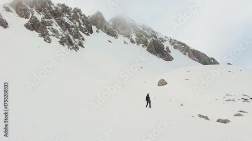 Hiker trekking alone through snowy mountain slopes surrounded by rocky peaks and mist.

