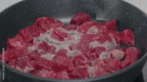 Chunks of raw beef are sizzling in a dark pan, with steam and bubbles rising from the meat as it cooks