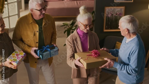 View from above of three senior friends giving presents to happy 70-year-old lady, kissing her on cheek and applauding while celebrating birthday in home setting