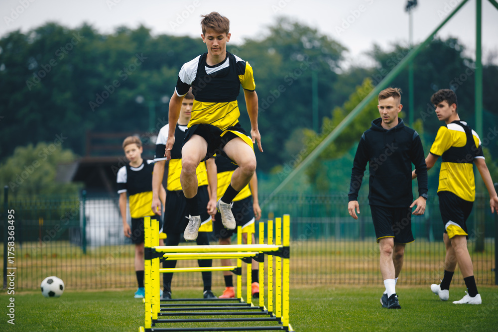 Fototapeta premium Player kick soccer ball to coach and ladder skipping. Teenagers on soccer training camp. Boys practice football with young coaches. Junior level athletes improving soccer skills on outdoor training