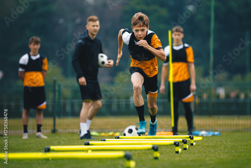 Teenage Boy at Training Drill With Young Soccer Coach. Youth Football Player Jumping Over Row of Hurdles and Running Fast at Sports Practice Field. Sports Strength and Agility Training For Footballers