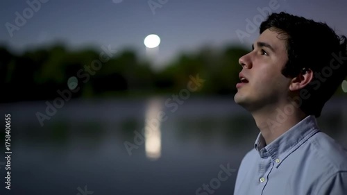 A young man with a look of wonder gazes up at the moon by a tranquil lake at night.