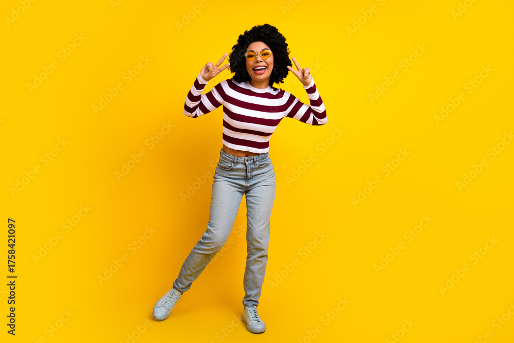Fototapeta premium Funny young woman in casual striped top posing against vibrant yellow backdrop with a joyful peace hand gesture