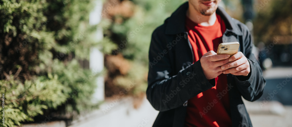 Fototapeta premium A young male outdoors interacting with his smartphone, casually dressed in a red shirt and black jacket with a cheerful expression.