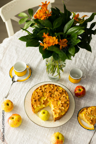 Homemade apple pie with custard cream on lace tablecloth, bouquet of red flowers and fresh apples