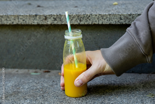 A person in a brown hoodie sets down a glass bottle filled with vibrant orange smoothie, possibly mango or carrot-based, featuring a mint-striped paper straw, on a stone step