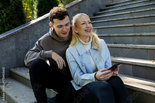 Young man and woman sit side by side on sunlit stone stairs, sharing a joyful moment as the woman smiles brightly while holding a smartphone, and the man looks on warmly