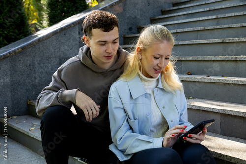 A young woman in a light blue jacket smiles at her phone while sitting on sunlit steps, as a young man in a brown hoodie leans in beside her, sharing the experience with interest
