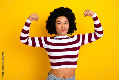 Confident woman flexing arms in striped top against vibrant yellow backdrop showcasing style and strength
