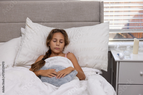 A young girl lying in bed holding a hot water bottle on her stomach to relieve pain.Symbolof illness, abdominal pain, home ca, and recovery.