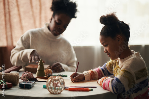 Black girl drawing with colored pencils at table while Black woman wrapping gifts in background, both engaging in Christmas holiday activities with festive decorations and craft supplies