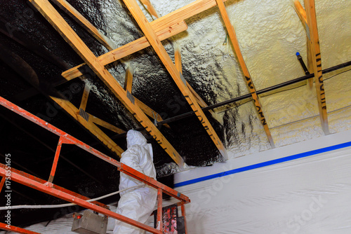 Fotografie Contractor in protective gear applies refractory paint insulation using spray gun to wooden ceiling beams at construction site