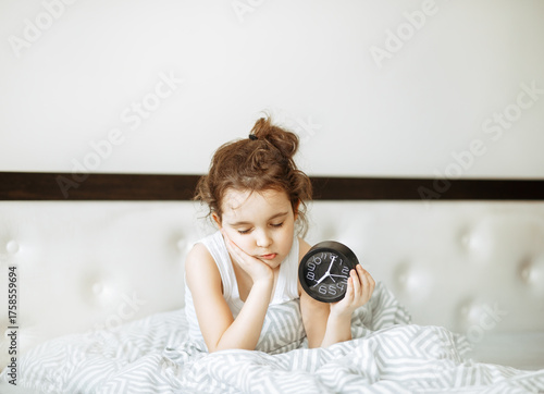 Cute little kid sleepy girl sitting on bed waking up in morning and holding alarm clock.