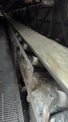 A dynamic, close-up shot captures a heavy industrial conveyor belt transporting a high volume of crushed material, such as cement or limestone, at great speed through a dark factory interior. The stee