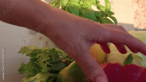On the table are red pepper, a head of fresh cabbage, basil, and celery. A hand is taking a lemon and placing it down. Close-up shot.